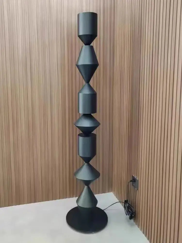 Tall black geometric sculpture on a white surface with a wooden wall background