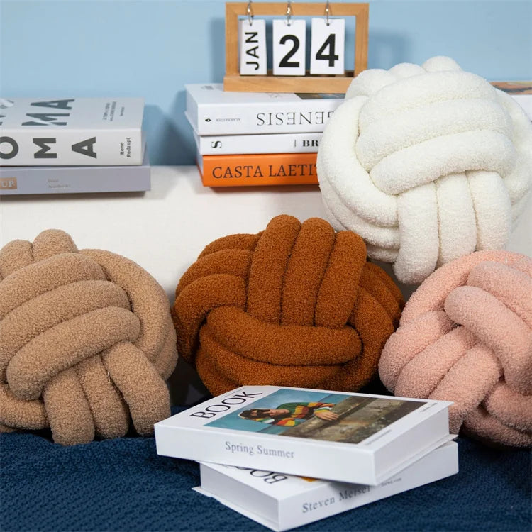 Decorative knot pillows in various colors on a surface with books and a calendar in the background.