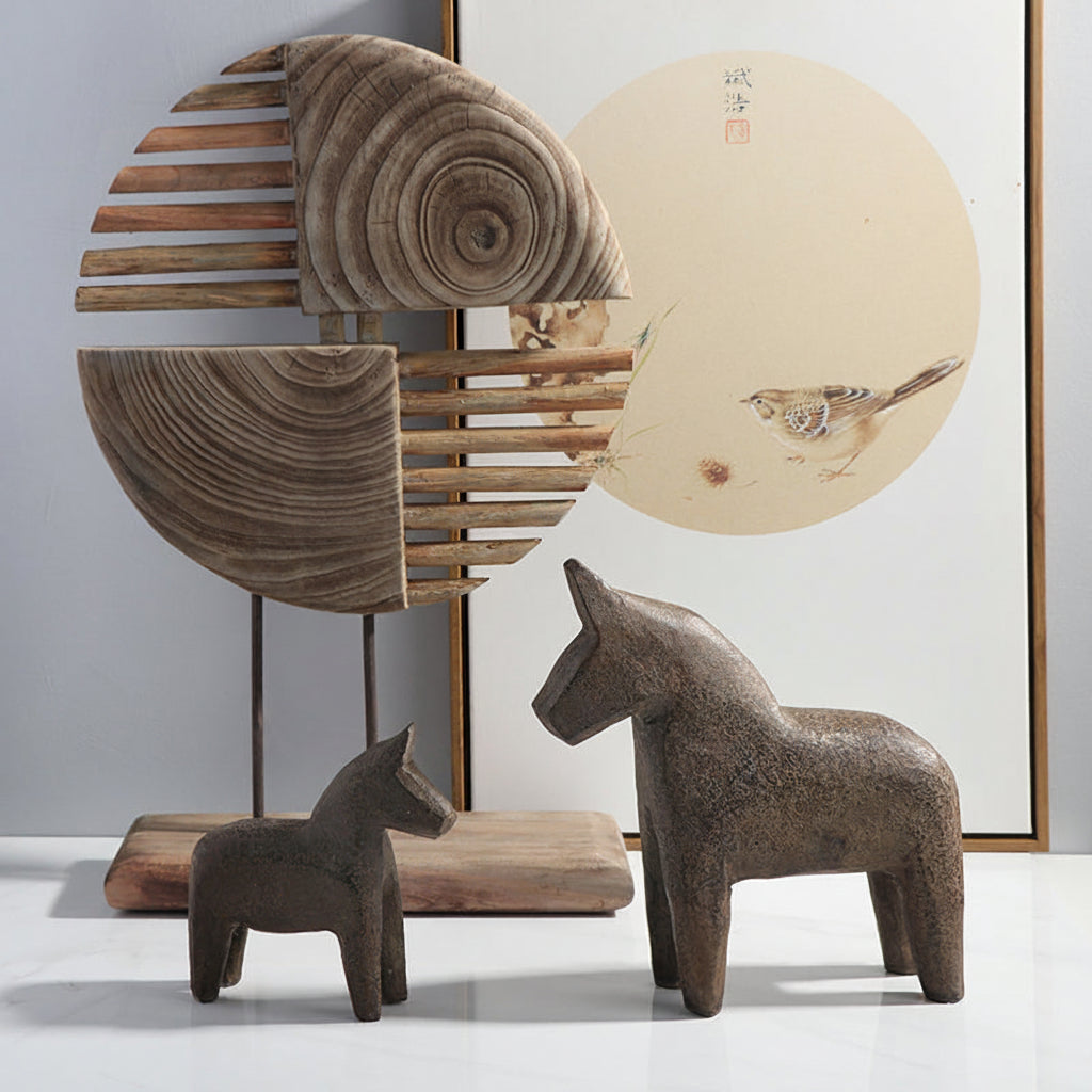 Two brown ceramic horse sculptures on a white surface with a wooden sculpture and framed artwork in the background.