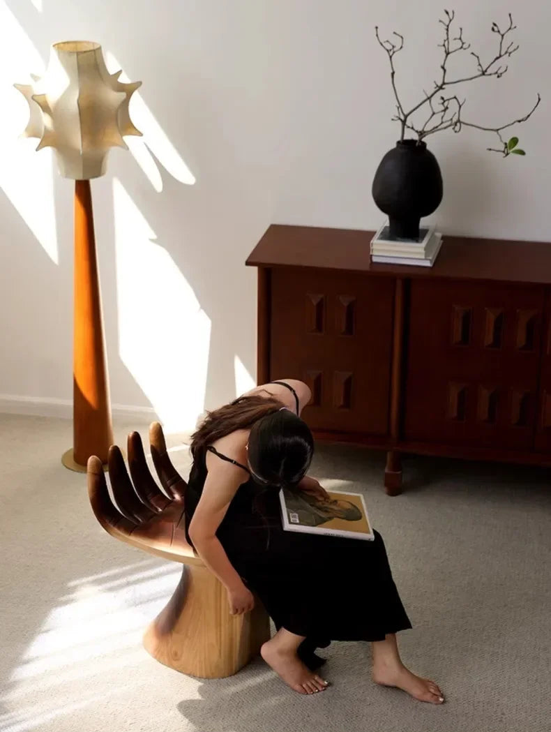 Woman sitting on a wooden chair reading a book in a room with a lamp and cabinet.