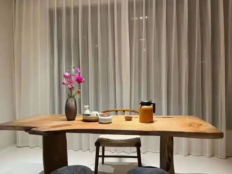 Wooden table with a vase of flowers and a teapot in a room with light gray curtains.
