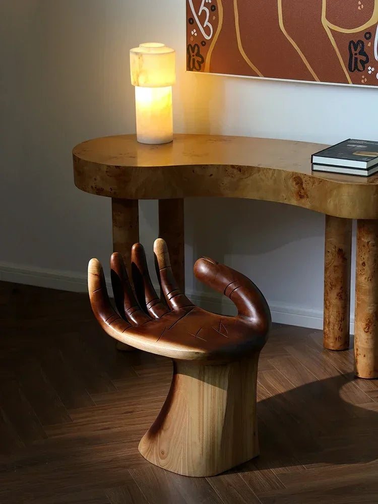 Wooden hand-shaped chair in a room with a lamp and books on a table.