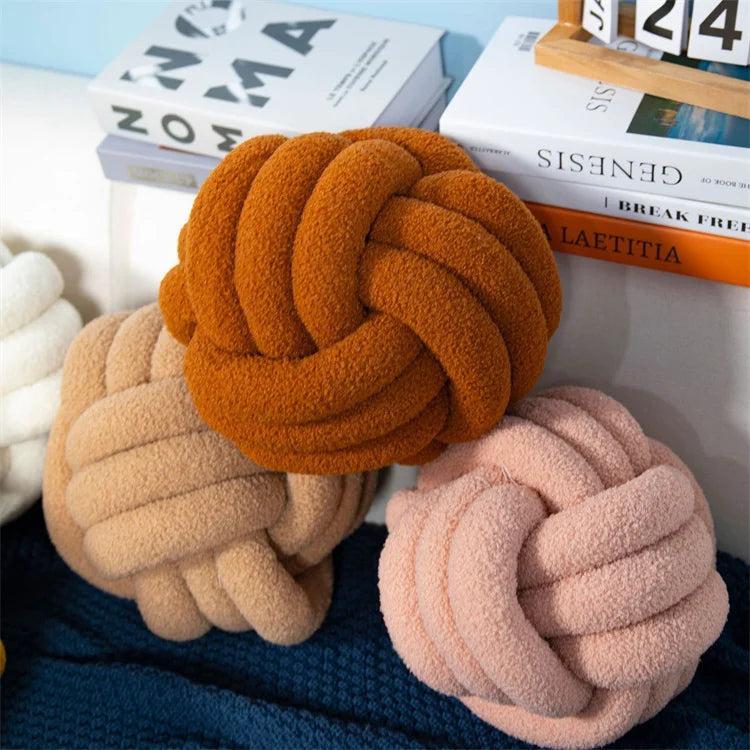 Three knotted plush toys in orange, beige, and pink on a surface with books in the background.