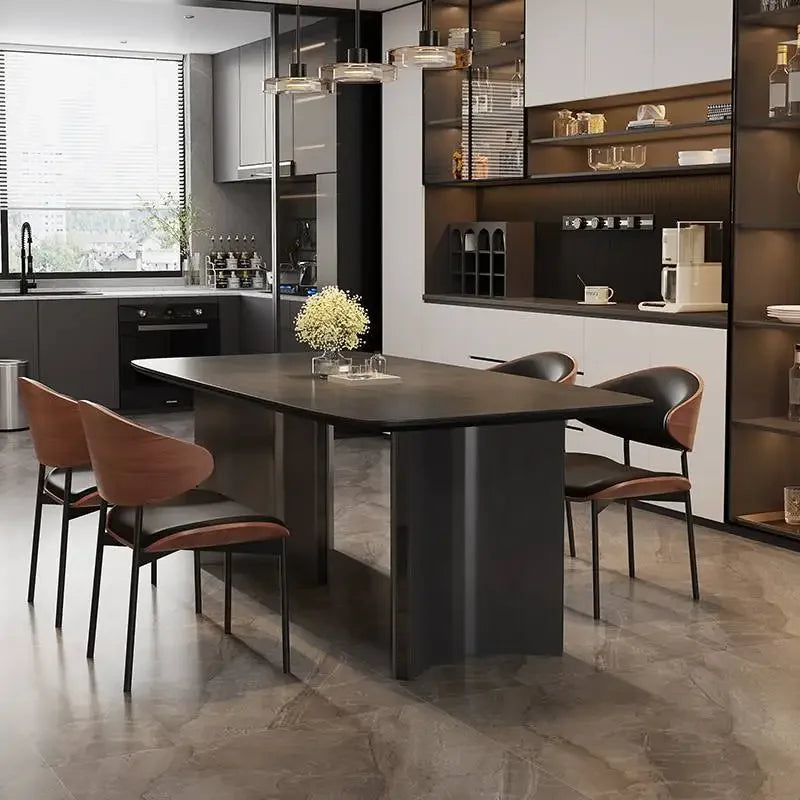 A modern dining room setup featuring a dark rectangular table with a thick pedestal base, surrounded by four contemporary chairs with brown leather seats and black metal frames. The scene is set in a contemporary kitchen with dark cabinets, open shelving, and a tiled floor.