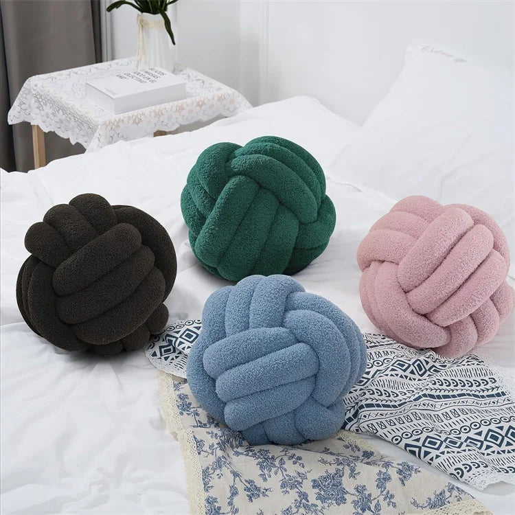 Four knot-shaped cushions in black, green, blue, and pink on a bed.