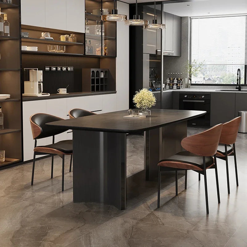 A modern dining room setup featuring a dark rectangular table with a central pedestal base, surrounded by four contemporary chairs with brown leather and black metal frames. The setting is a stylish, contemporary kitchen with dark cabinets, open shelving, and a large window.