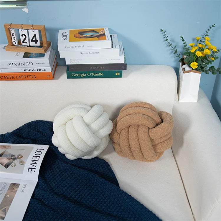 Decorative knot pillows on a sofa with books and a plant in the background.