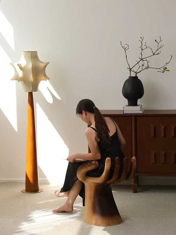 Woman sitting on a unique wooden chair in a modern interior setting with a lamp and vase.