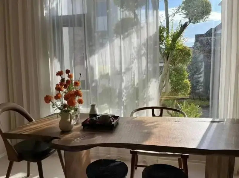 Dining area with a wooden table, chairs, and a vase of flowers in front of a large window.